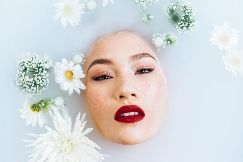 Imagem de um rosto oriental. Sobre ele algumas flores brancas, enfeitando. É uma modelo com pele branca, olhos pintados e boca com um lindo batom na cor vermelho.