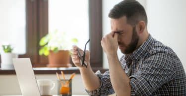 Homem sentado em mesa de trabalho com mão nos olhos e expressão de estress