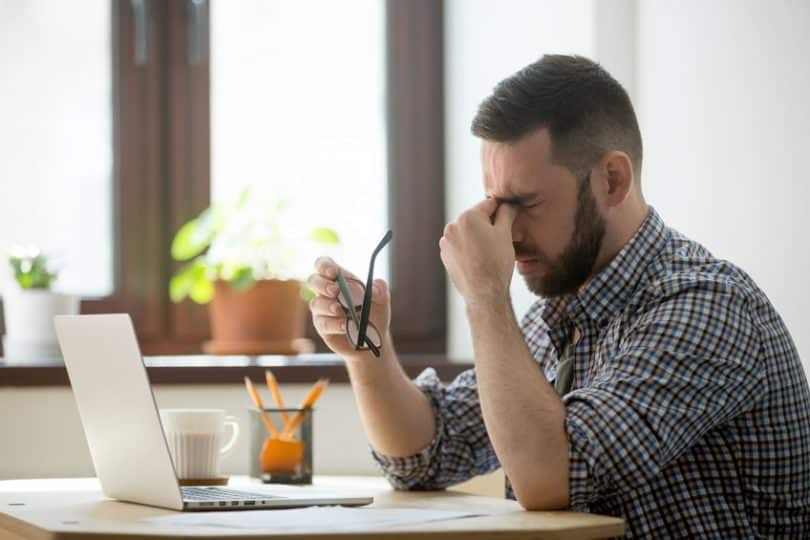 Homem sentado em mesa de trabalho com mão nos olhos e expressão de estress
