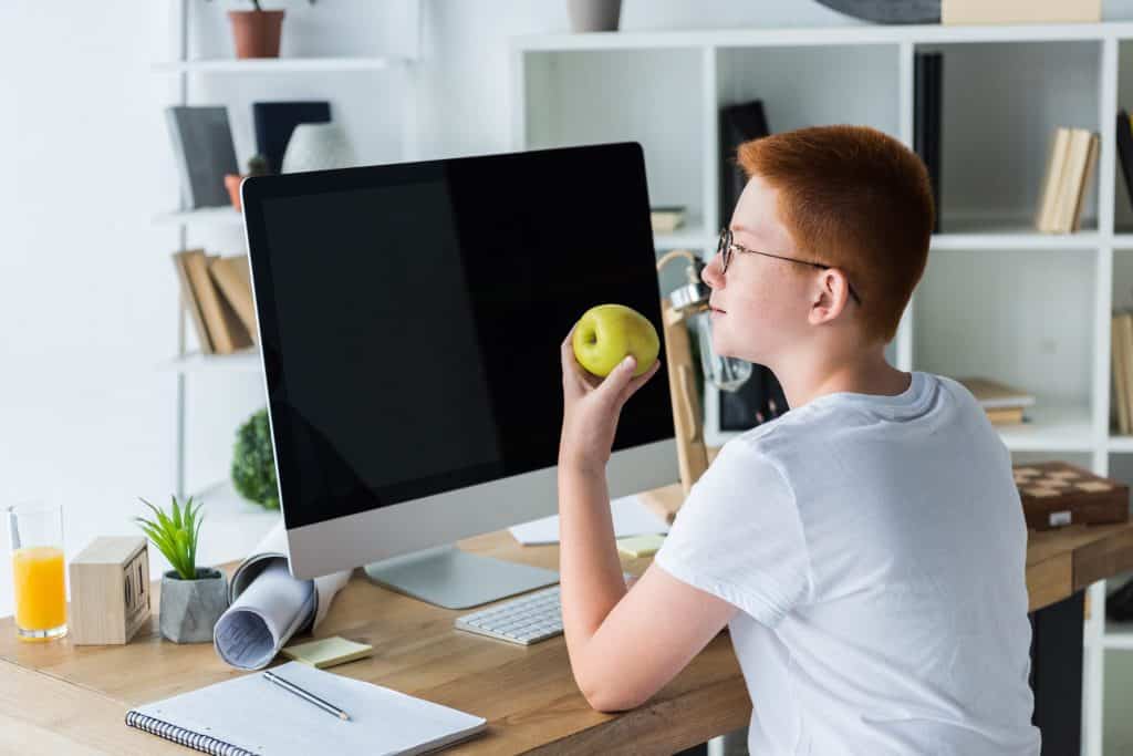 Imagem de um jovem garoto de cabelo curto e ruivo, vestindo uma camiseta básica na cor branca. Ele está sentado de frente para o seu computador e deu uma pausa nas redes sociais enquanto degusta uma linda e saborosa maçã verde.