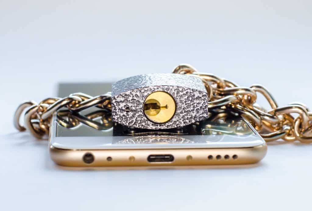 Imagem de um celular dourado envolto de uma corrente com cadeado. Ele está disposto sobre uma mesa branca.