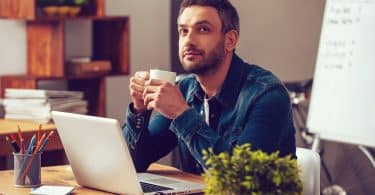 Homem branco de barba castanha, sentado em frente a uma mesa de escritório, enquanto segura uma caneca branca.