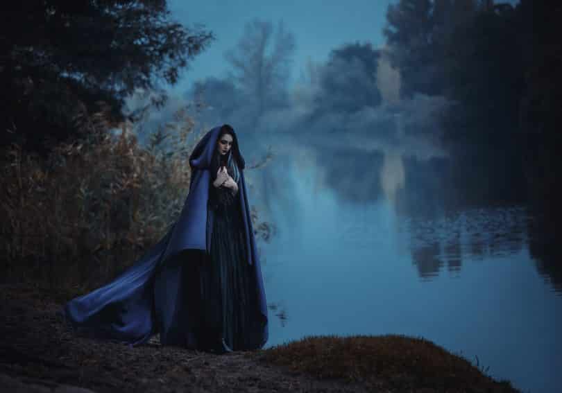 Mulher branca com roupas e capa escuras, ao lado de um lago.