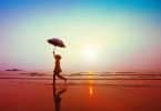 Mulher correndo na areia da praia segurando guarda-chuva.