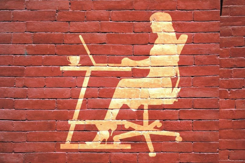 Imagem artística da silhueta de uma mulher empreendedora sentada frente à uma mesa com o seu computador. A imagem está refletida em uma parede feita de tijolos vermelhos.