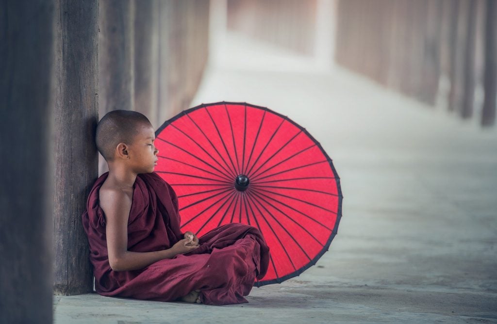 Imagem de um jovem budista sentado e encostado em um tronco de árvore. Ao lado dele um guarda-chuva vermelhor aberto. Ele está meditando.