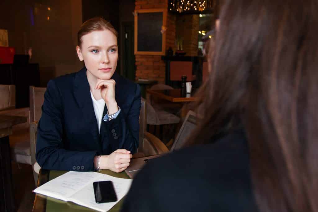 Imagem de duas mulheres empreendedoras fazendo uma reunião de negócios em um restaurante. Elas estão usando camisa social e um terno escuro. Uma delas aparece estar apreensiva.
