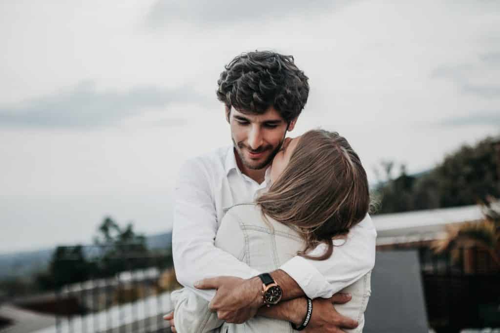 Homem e mulher brancos, vestindo roupas brancas, se abraçando.