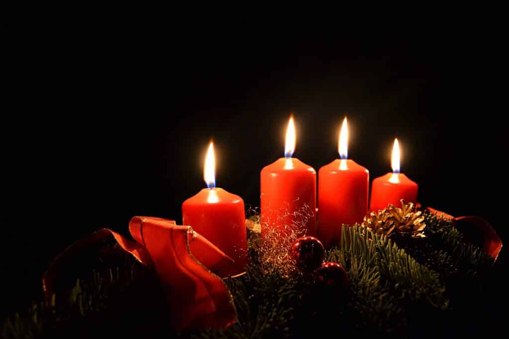 Imagem de um lindo arranjo composto por quatro velas na cor vermelha. Elas estão acesas e dispostas sobre um suporte decorado com ramos de árvores na cor verde, pinhas, bolas vermelhas e laço de fita tamém na cor vermelha,