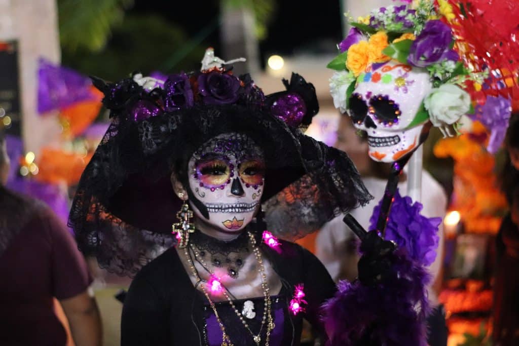 Imagem de uma moça fantasiada de La Catrina, a Deusa da Morte. Ela usa um vestido preto e um chapéu de renda preto. Seu rosto está pintado de caveira. Usa brincos e colares dourados bem grandes.