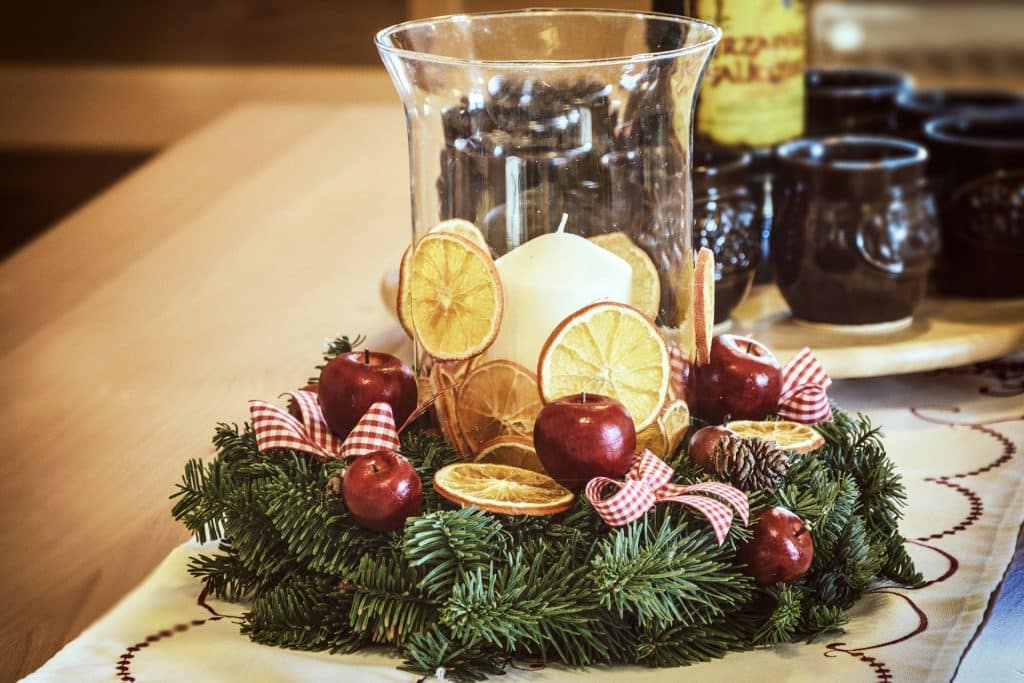 Imagem de um lindo arranjo de mesa natalino decorado com uma guirlanda feita de folhagem verde, maçãs inteiras, rodelas de laranja, fitas branca e vermelha e pinhas.
