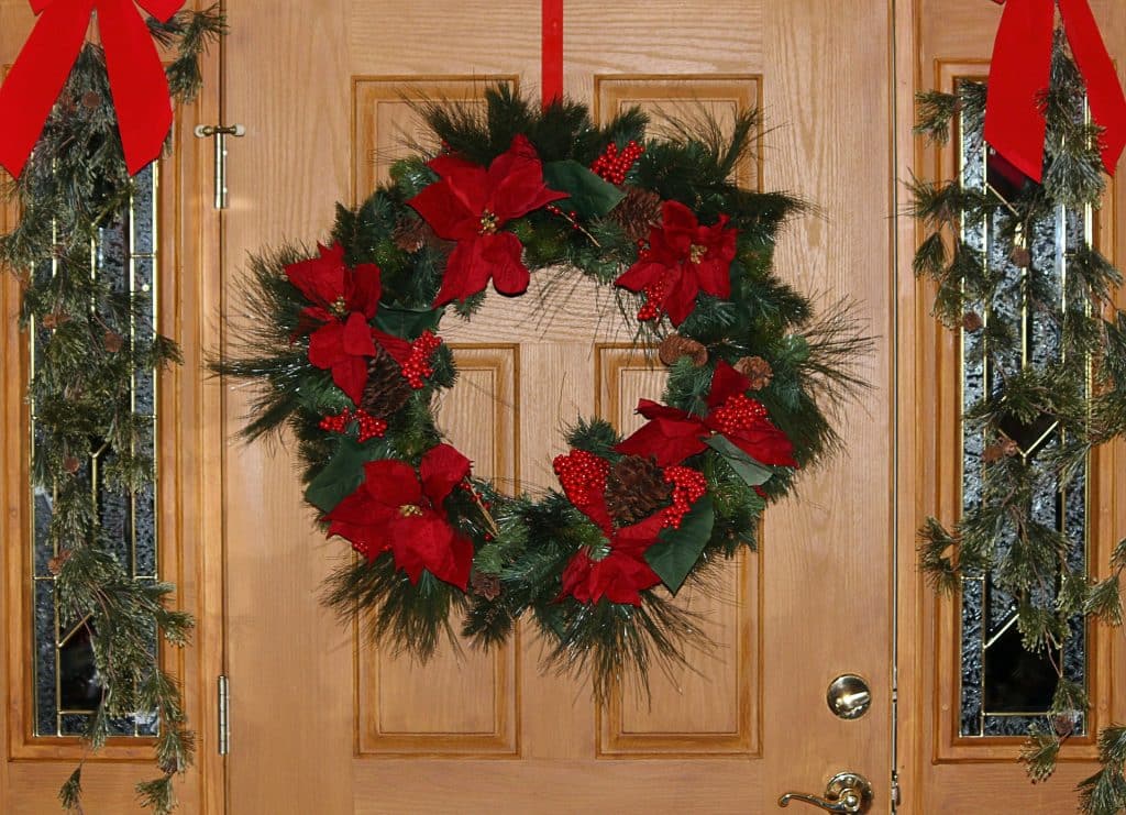 Imagem de uma linda e grande guirlanda de Natal pendurada na porta de entrada de uma casa. A guirlanda é feita de folhas verdes decoradas com flores e folhagens vermelha.