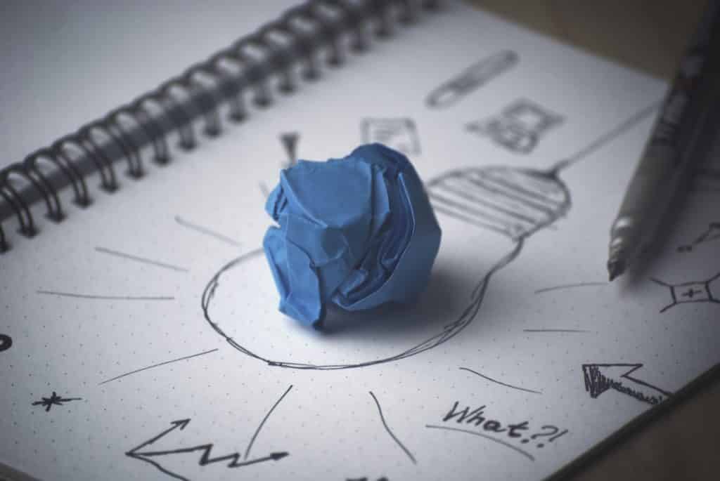 Imagem de um caderno com o desenho de uma lâmpada, setas, gráficos e sobre ele um papel post-it na cor azul, amassado.