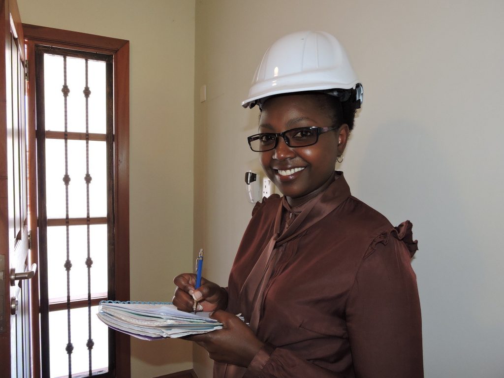 Imagem de uma mulher negra trabalhando. Ela usa um capacete branco, óculos de grau e segura em suas mãos uma caneta e um caderno.