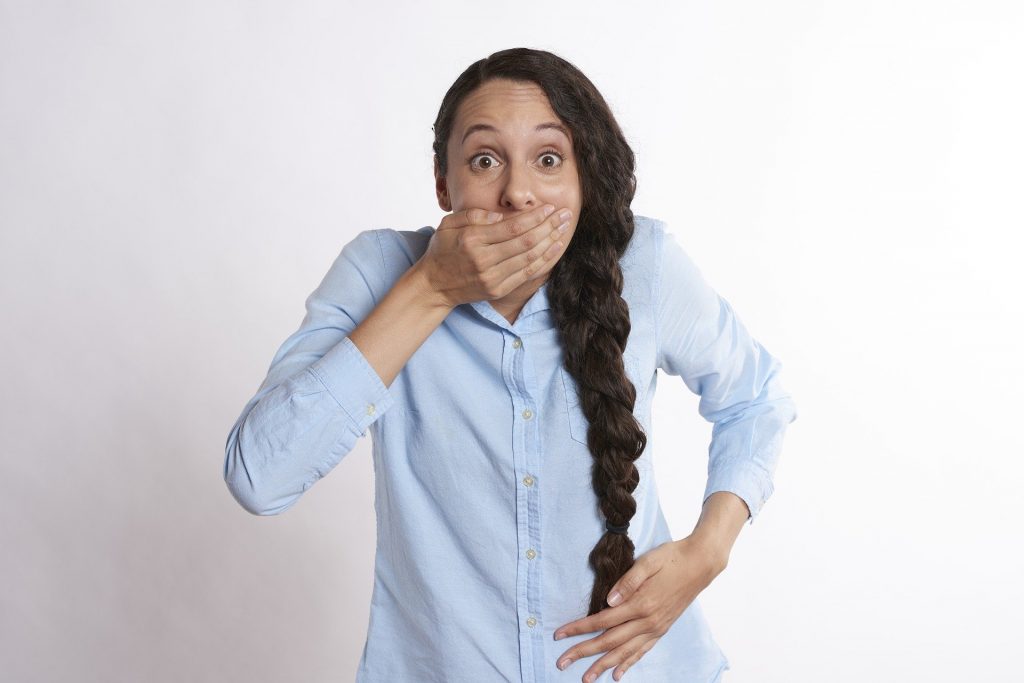 Imagem de uma mulher vestindo uma camisa azul clara. Ela usa uma trança longa em seus cabelos castanhos e está com uma das mãos sobre a boca.
