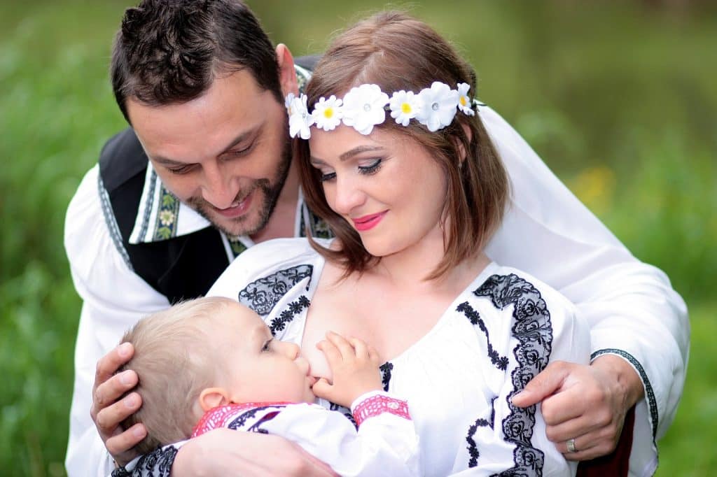 Imagem de um casal. A mulher está amamentando o seu filho. Ela usa um adereço florido na cabeça e ambos vestem uma camisa branca bordada com renda preta.