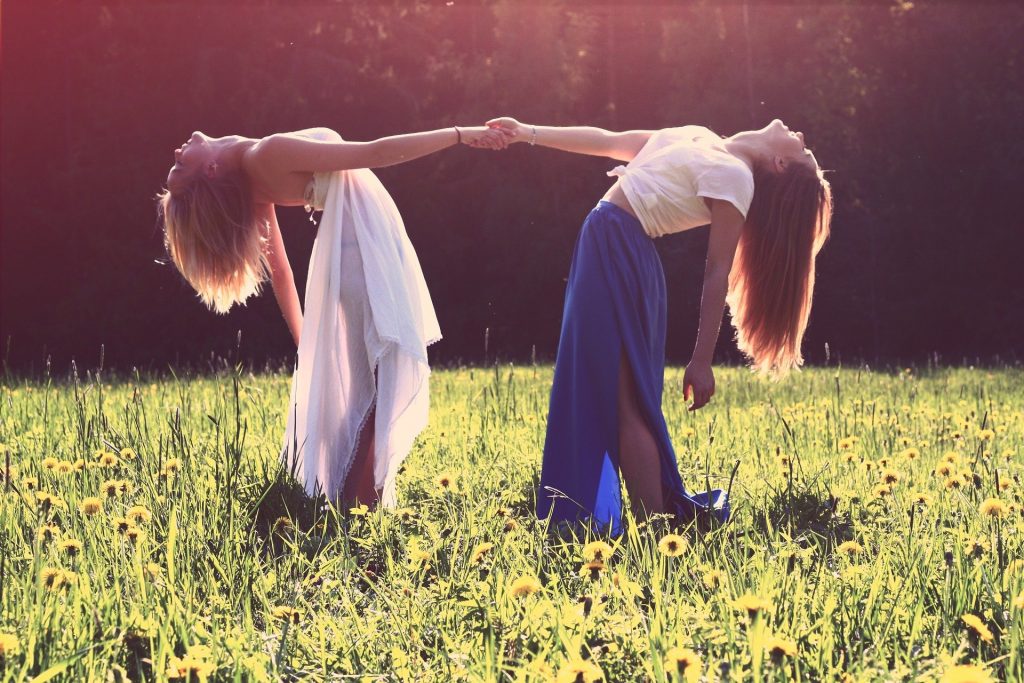 Imagem de duas garotas dando as mãos. Elas estão em um lindo jardim florido. Usam saias longas e estão felizes, praticando a gentileza entre elas.
