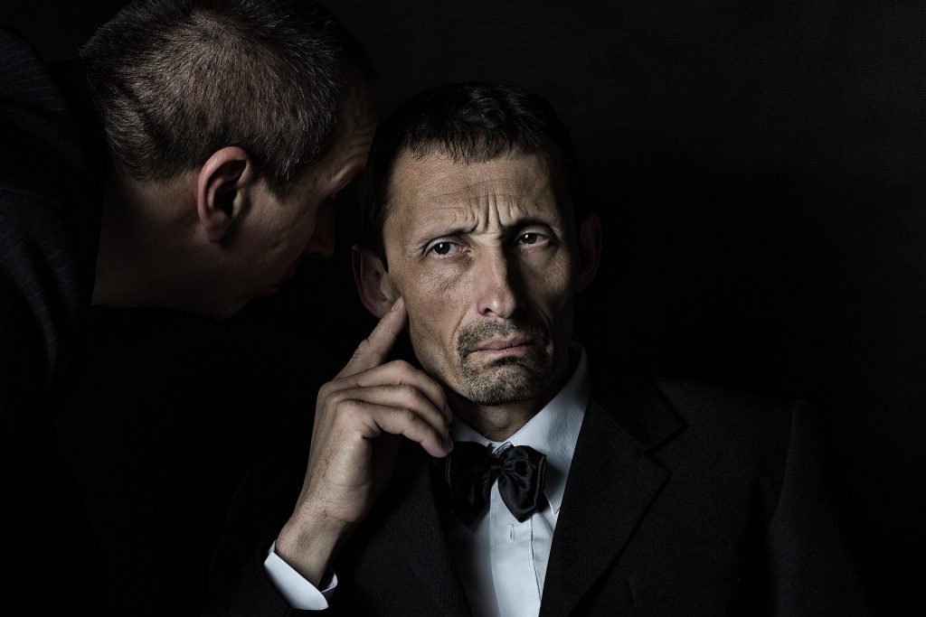 Imagem de dois senhores. Um está sentando e ouvindo o que o outro está dizendo em seu ouvido.