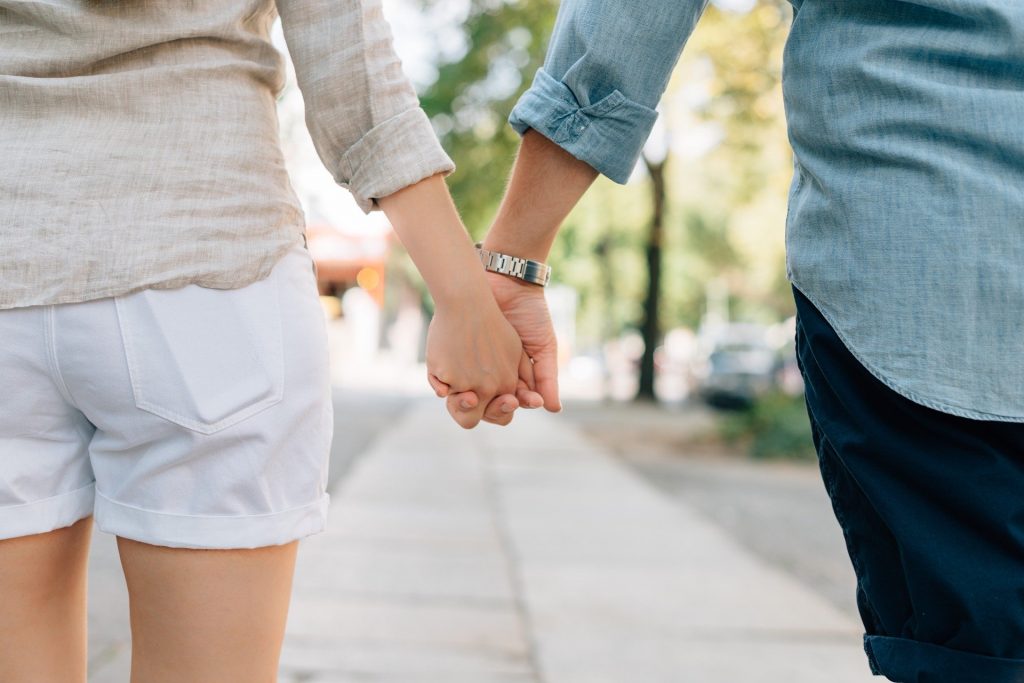 Imagem de um casal de costas, caminhando de mãos dadas. Ela usa um shorts branco e uma camisa bege e ele uma bermuda azul marinho e uma camisa azul clara. Ele estão planejando a vida para 2021.