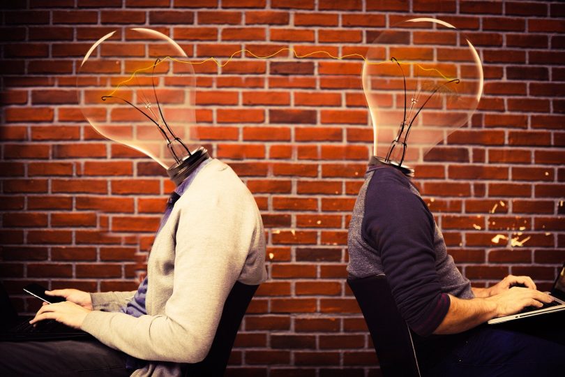 Imagem de dois homens vestidos de social. Eles estão sentados um de costas para o outro e ambos estão mexendo no celular e no computador. A cabeça dos dois é representada por uma lâmpada que configura a ideia, a criatividade.