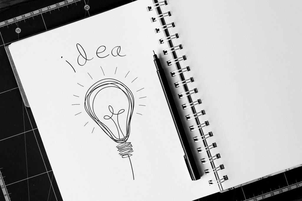 Imagem em preto e branco de um caderno de desenho grande. Nele está desenhado uma lâmpada e sobre ela está escrito a palavra "idea". Ao lado da lâmpada desenhada uma caneta de desenho na cor preta.