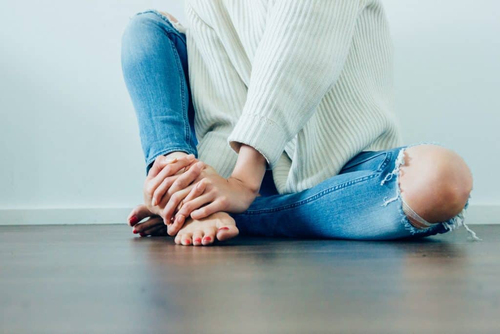 Mulher branca usando suéter branco e calça jeans rasgada, sentada no chão e com as mãos em um dos pés.