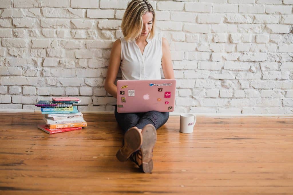 Imagem de uma mulher sentada no chão com piso de madeira ela está segurando um notebook e trabalhado com redes sociais.