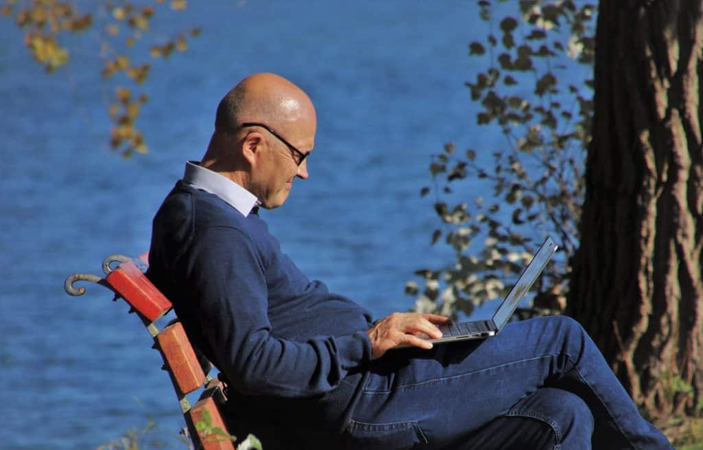 Imagem de um senhor sentado em um banco de mandeira. Ele está mexendo no notebook. Ele usa blusa de frio e calça jeans. Ao fundo a imagem do mar azul.