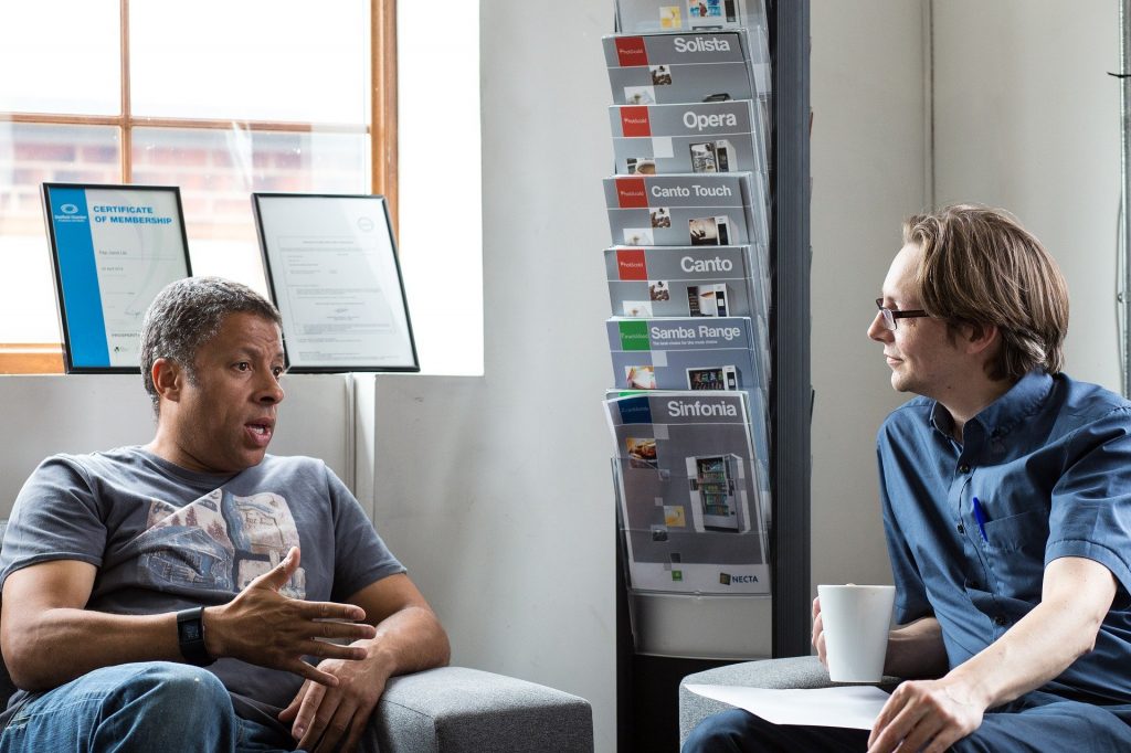 Imagem de uma sala de trabalho e nela dois homens conversando. Um deles segura uma xícara branca de café e o outro apenas gesticula com uma de suas mãos.