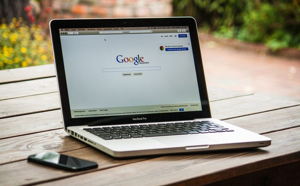 Imagem de um notebook aberto na página do google. O computador está sobre uma mesa de madeira e ao lado dele um aparelho de telefone celular.