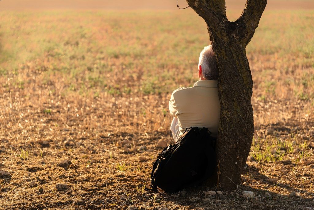 Imagem de um campo gramado e de um homem sentado e encostado em uma árvore. Ele está só e pensativo.