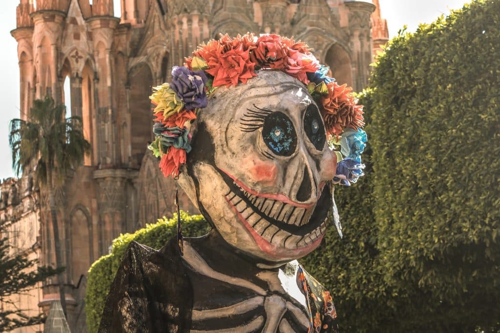 Imagem da Deusa da Morte, La Catrina. Ela usa uma máscara de caveira e um arco de flores em sua cabeça. Ela está em um jardim e ao fundo um castelo antigo.
