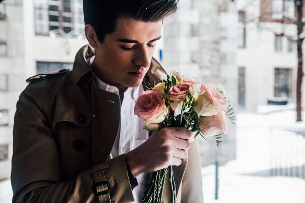 Imagem de um modelo masculino usando uma jaqueta de frio marrom e uma camisa branca. Em uma das mãos ele segura um ramalhete de rosas.