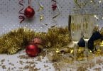 Imagem de uma decoração bem festiva para celebrar as festas de final de ano. A decoração é composta por: festão dourado, bolas vermelhas e douradas, taças com champanhe, serpentina.