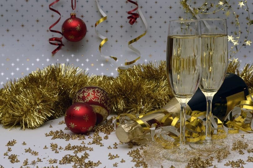 Imagem de uma decoração bem festiva para celebrar as festas de final de ano. A decoração é composta por: festão dourado, bolas vermelhas e douradas, taças com champanhe, serpentina.