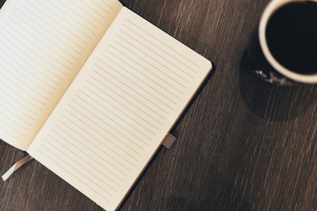 Imagem de um caderno aberto com folhas em branco. Ao lado uma xícara com café. O caderno está sobre uma mesa de madeira.