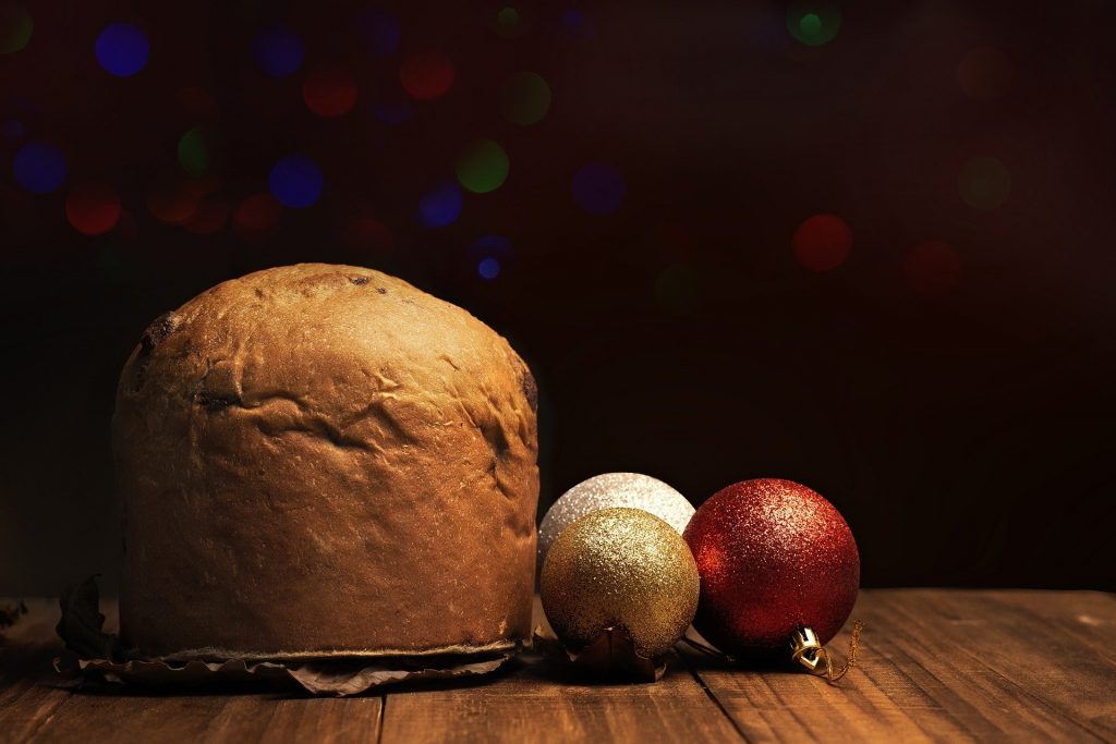 Imagem de um panetone inteiro disposto sobre uma mesa de madeira e ao lado dele três bolas de enfeites de Natal nas cores: vermelho, branco e dourado.
