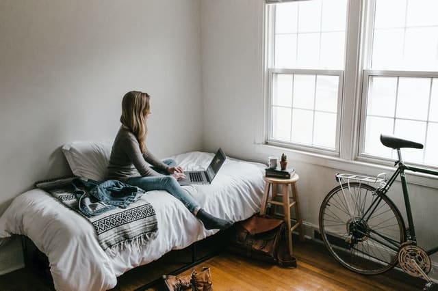 Mulher sentada na cama mexendo no notebook com quarto arrumado