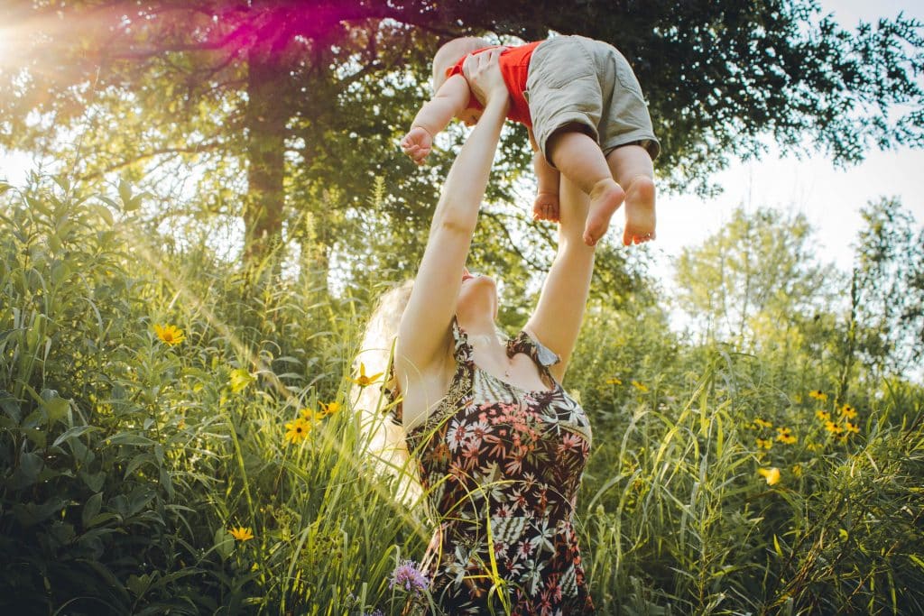 Mãe levantando filho com sol refletindo ao fundo em campo de mato e flores