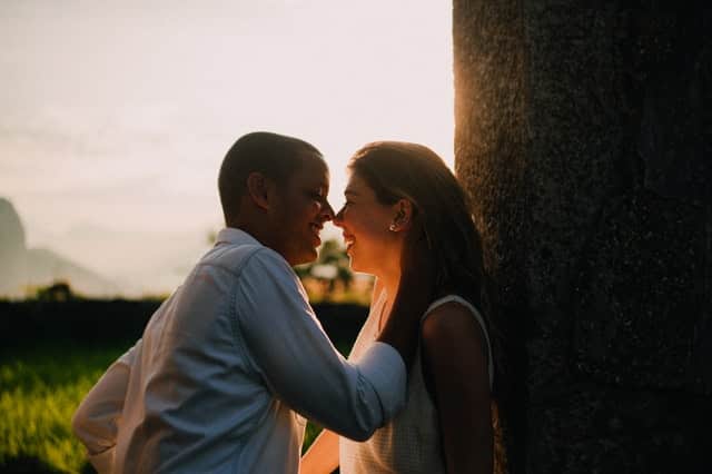 Casal sorrindo com homem com mão na nuca de mulher e sol ao fundo refletindo levemente