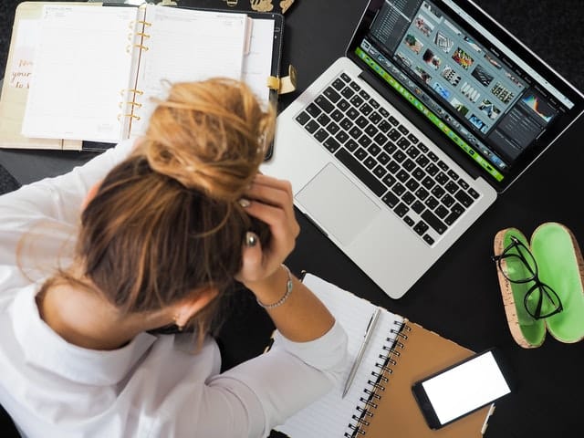 Mulher em mesa de trabalho com notebook, óculos, caderno e celular com cabeça abaixada em sinal de cansaço