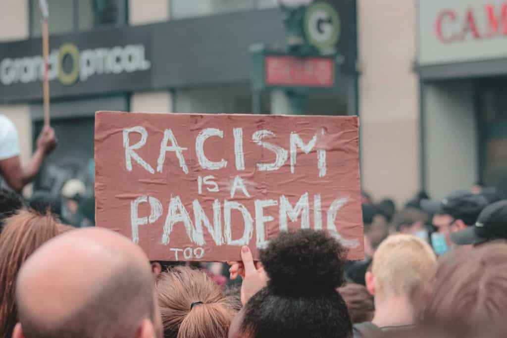 Manisfestação na rua com uma mulher segurando um cartaz escrito "Racismo é uma pandemia também"