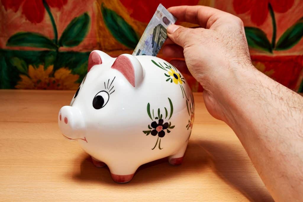 Imagem de um cofrinho de porcelana na cor branca feito em formato de um porquinho pintado à mão. A mão de um homem está ao fundo e ele está colocando uma cédula no cofrinho.