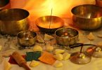 Imagem de vários elementos e itens para a prática do esoterismo. São vários tipos de bowls, incensos, pedras, velas, dispostos sobre uma mesa forrada com uma toalha branca.