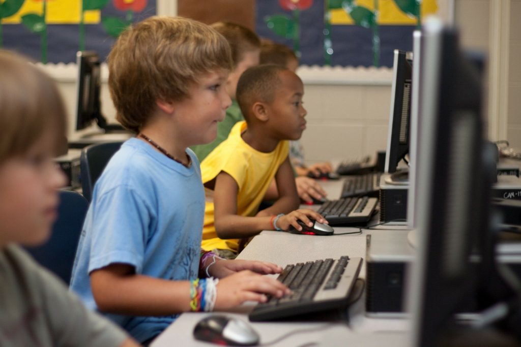 Imagem de uma sala cheia de crianças e com vários computadores. Muitas delas estão mexendo no computador.