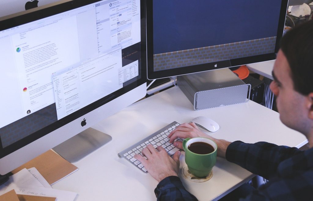 Imagem de um homem digitando. À sua frante uma caneca de café. Ele está trabalhando e usando duas telas para realizar as suas tarefas.
