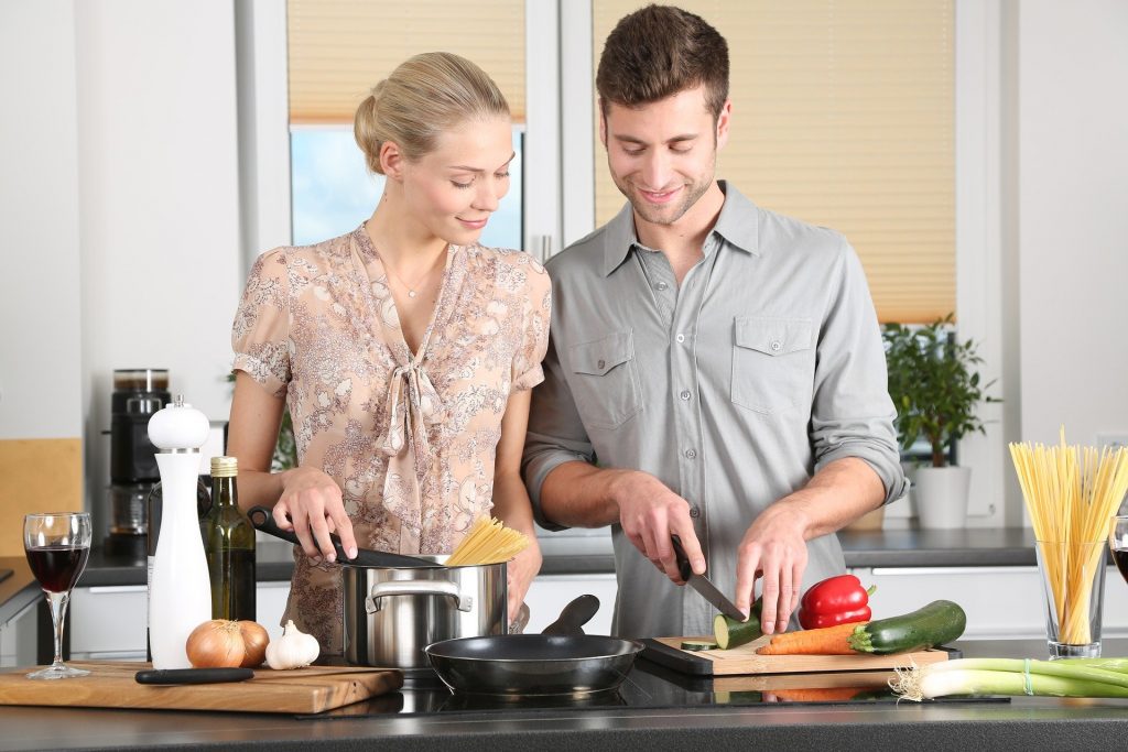 Mulher cozinhando ao lado do marido, ensinando ele a fazer um delicioso jantar. Ela está cozinhando uma massa de macarrão e ele cortando os legumes para uma bela salada.