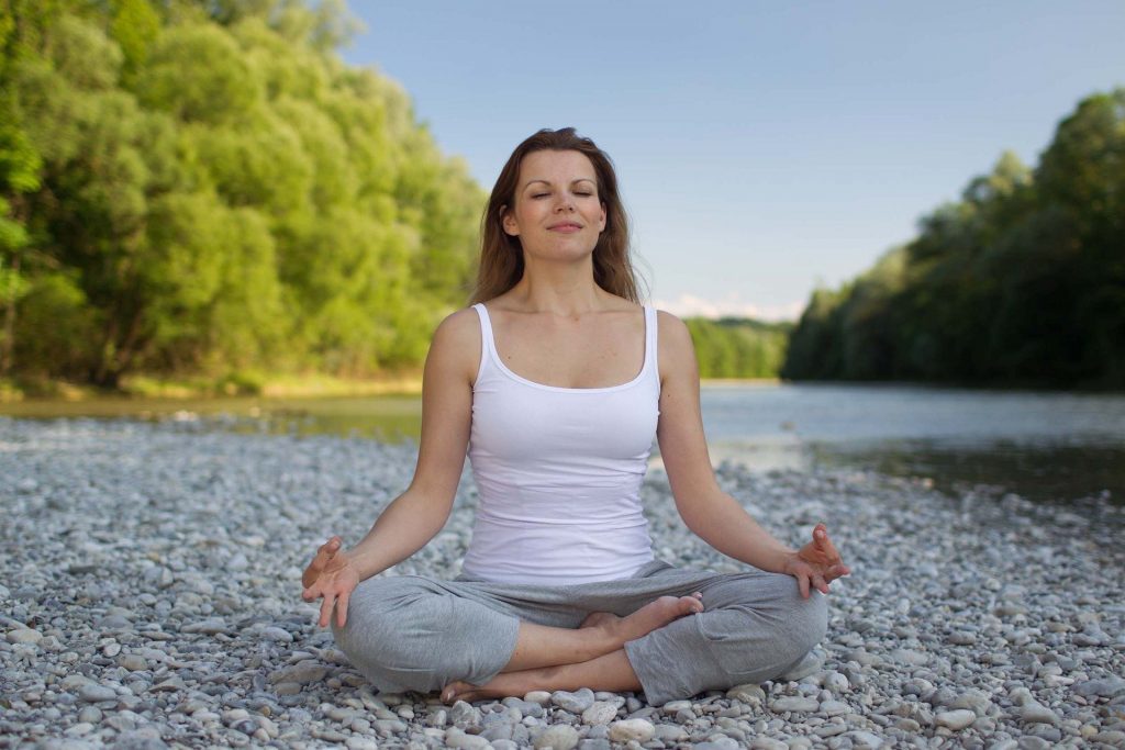 Imagem de uma mulher sentada em posição de yoga. Ela usa uma camiseta regata branca e uma calça cinza de moletom.
