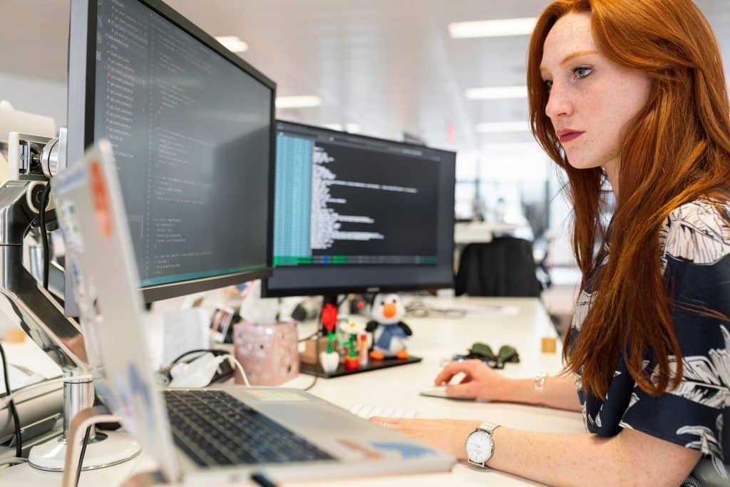 Imagem de uma mulher ruiva com cabelos longos. Ela está trabalhando sentada à frente de três computadores.