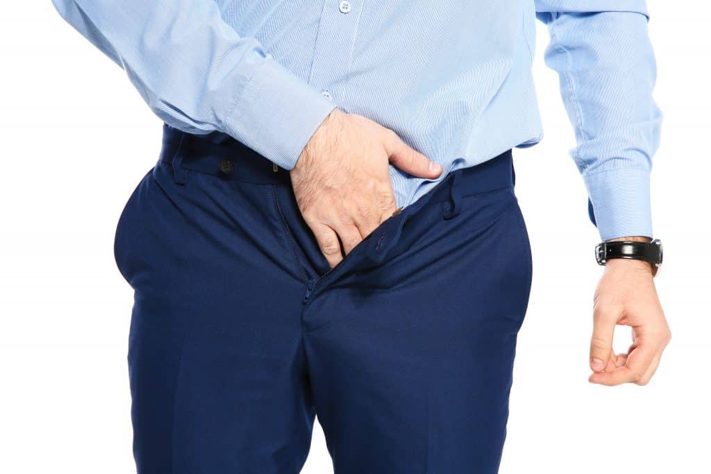 Imagem de um homem usando camisa e calça social na cor azul claro e escuro, respectivamente. Ele está com uma das mãos dentro da calça. Ele foi diagnosticado com uma infecção denominada candidíase masculina.
